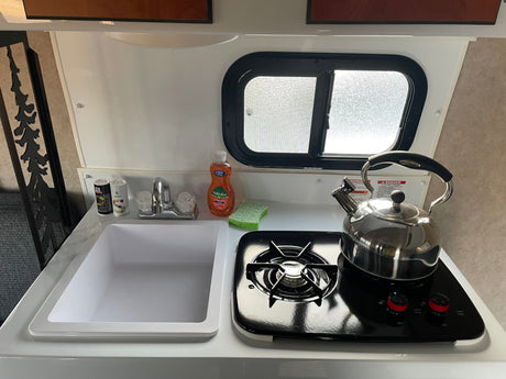 This RV rental offers a compact kitchen with a sink, single-burner stove, kettle, and window, perfect for cooking on the go.