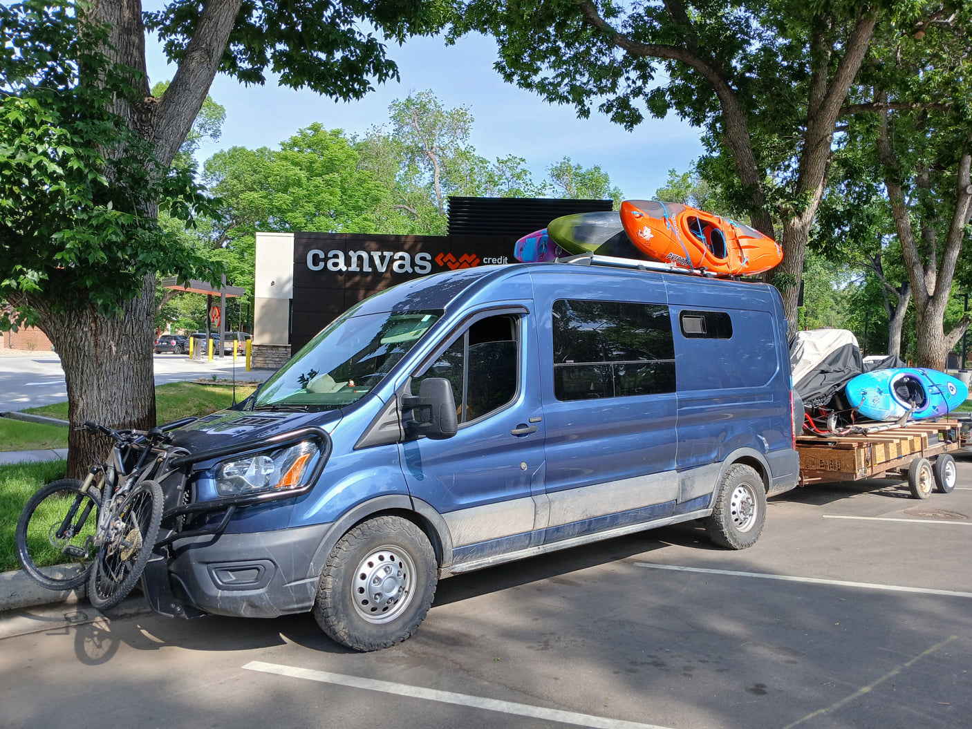 Blue RV rental van packed with kayaks & bikes, towing gear, ready for adventure under sunny trees.