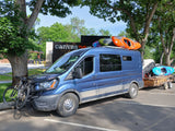 Blue RV rental van packed with kayaks & bikes, towing gear, ready for adventure under sunny trees.