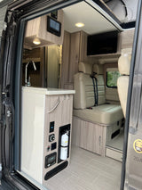 Interior of a clean rv rental van, showing a kitchenette, control panel, fire extinguisher, and passenger seating area.