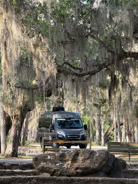 Grey RV rental van beneath majestic, moss-draped trees, with a park bench and rocks foregrounding a tranquil scene.