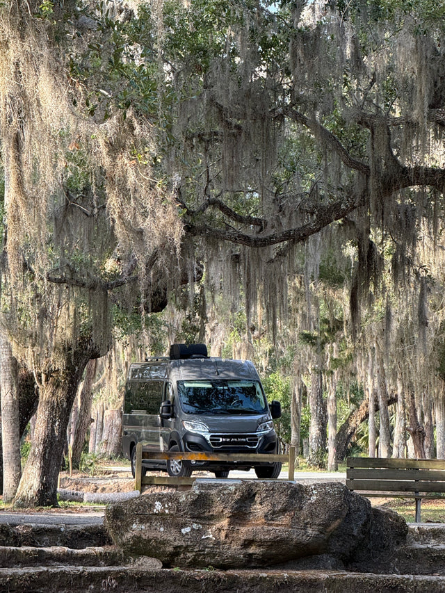 Grey RV rental van beneath majestic, moss-draped trees, with a park bench and rocks foregrounding a tranquil scene.