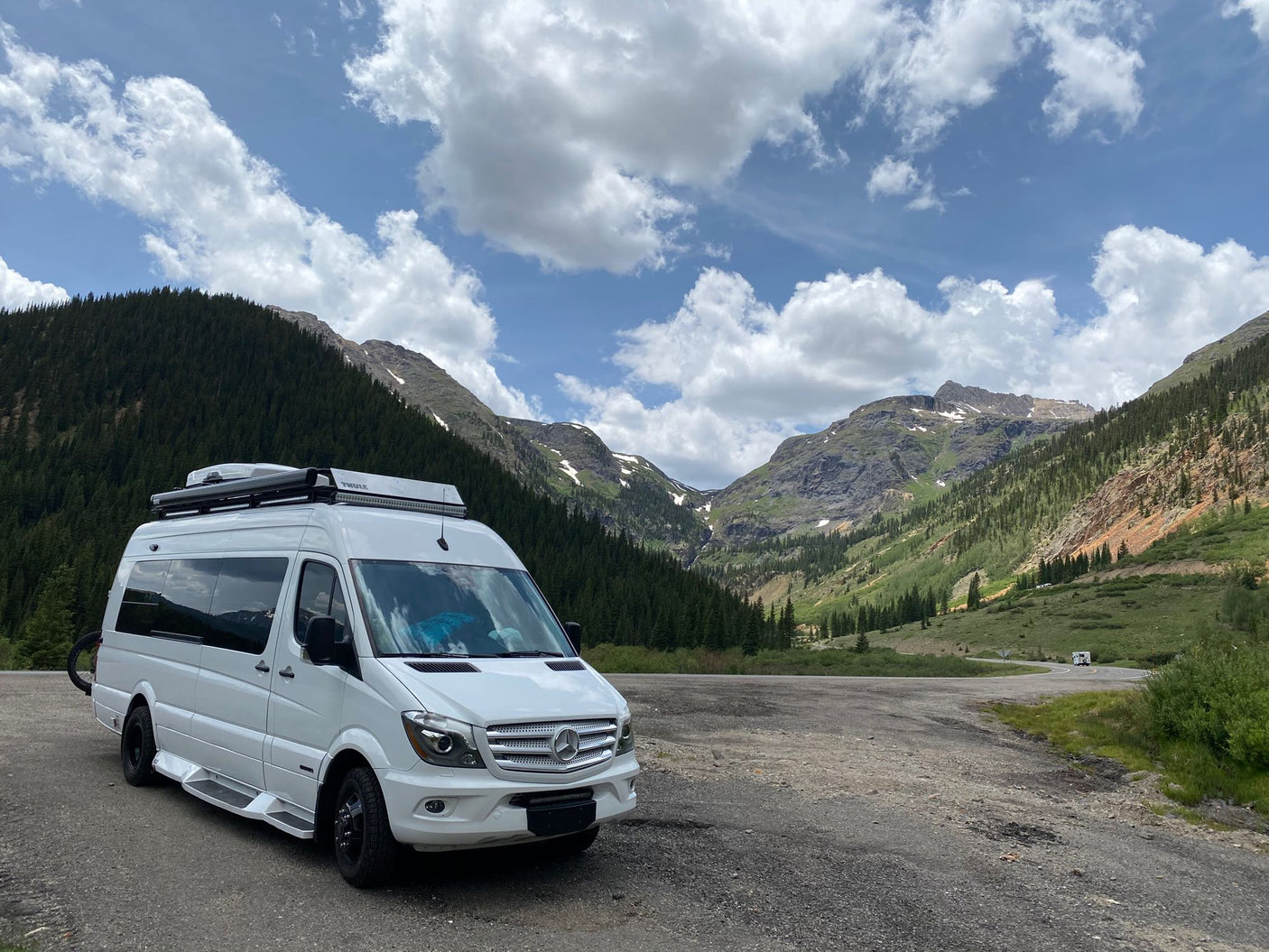 White Mercedes Sprinter rv rental in a stunning mountain valley. Green forests, snowy peaks & cloudy blue sky. Ideal for adventure.