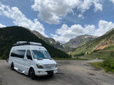 White Mercedes Sprinter rv rental in a stunning mountain valley. Green forests, snowy peaks & cloudy blue sky. Ideal for adventure.