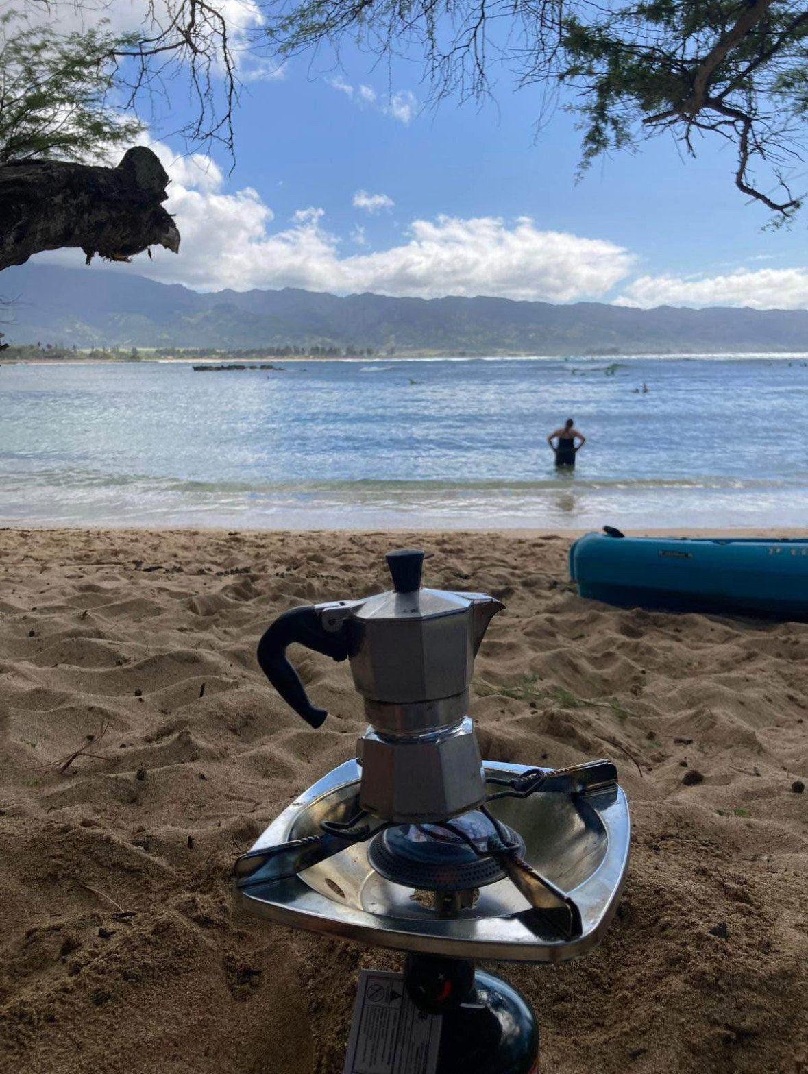 RV rental escape: Moka pot coffee brewing on a beach stove. Ocean waves, person, & mountains complete this outdoor scene.