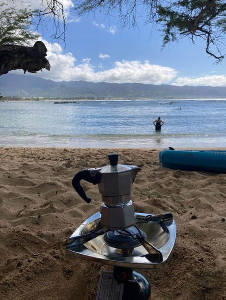 RV rental escape: Moka pot coffee brewing on a beach stove. Ocean waves, person, & mountains complete this outdoor scene.