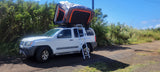 An RV rental experience: white Nissan Xterra SUV with open rooftop tent & ladder, parked by green bushes for camping.