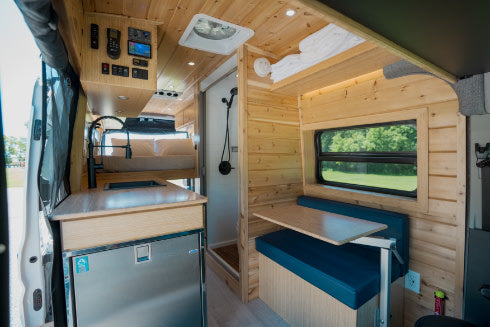 Inside a modern RV rental: a cozy camper van interior with a well-equipped kitchen, raised bed, and dining booth.