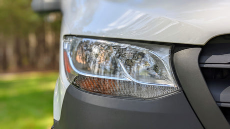 White RV rental van: close-up of its front right side, showing the headlight and black bumper. Outdoors in daylight.