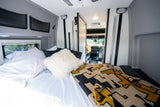 Cozy RV rental interior: a comfortable bed with pillows & a patterned blanket, looking into the camper van's front cabin.