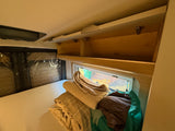 Inside an RV rental: A made-up bed with stacked linens and accessible open overhead storage in the cozy bedroom area.