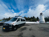 Silver Mercedes-Benz Sprinter RV rental parked at a historic lighthouse, nestled among evergreen trees under a cloudy blue sky.
