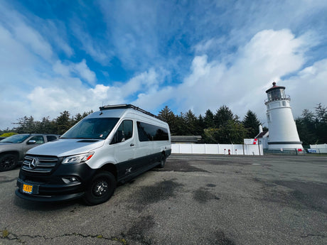 Silver Mercedes-Benz Sprinter RV rental parked at a historic lighthouse, nestled among evergreen trees under a cloudy blue sky.