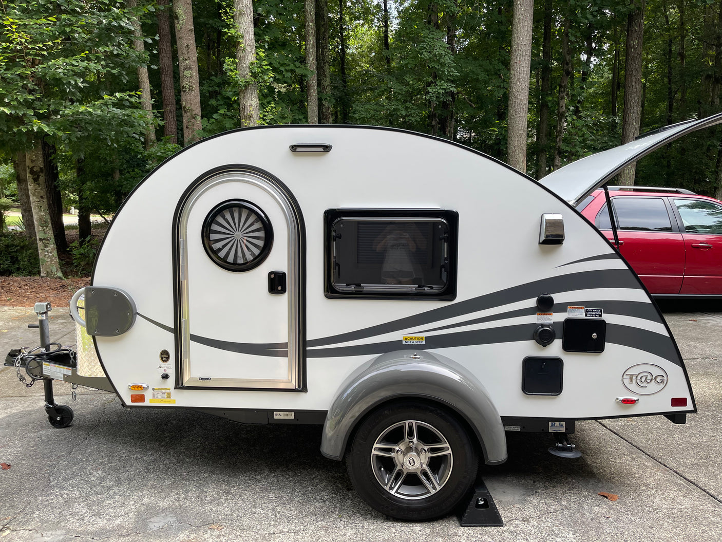 A white T@G teardrop camper, ideal for an rv rental or camping trip, is parked outdoors near trees.