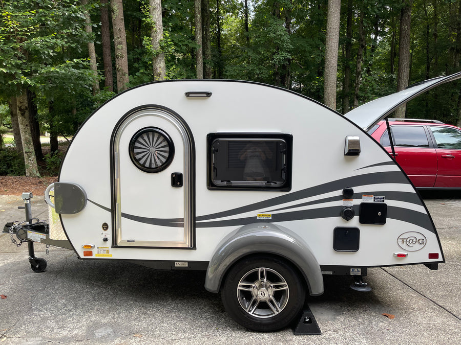 A white T@G teardrop camper, ideal for an rv rental or camping trip, is parked outdoors near trees.