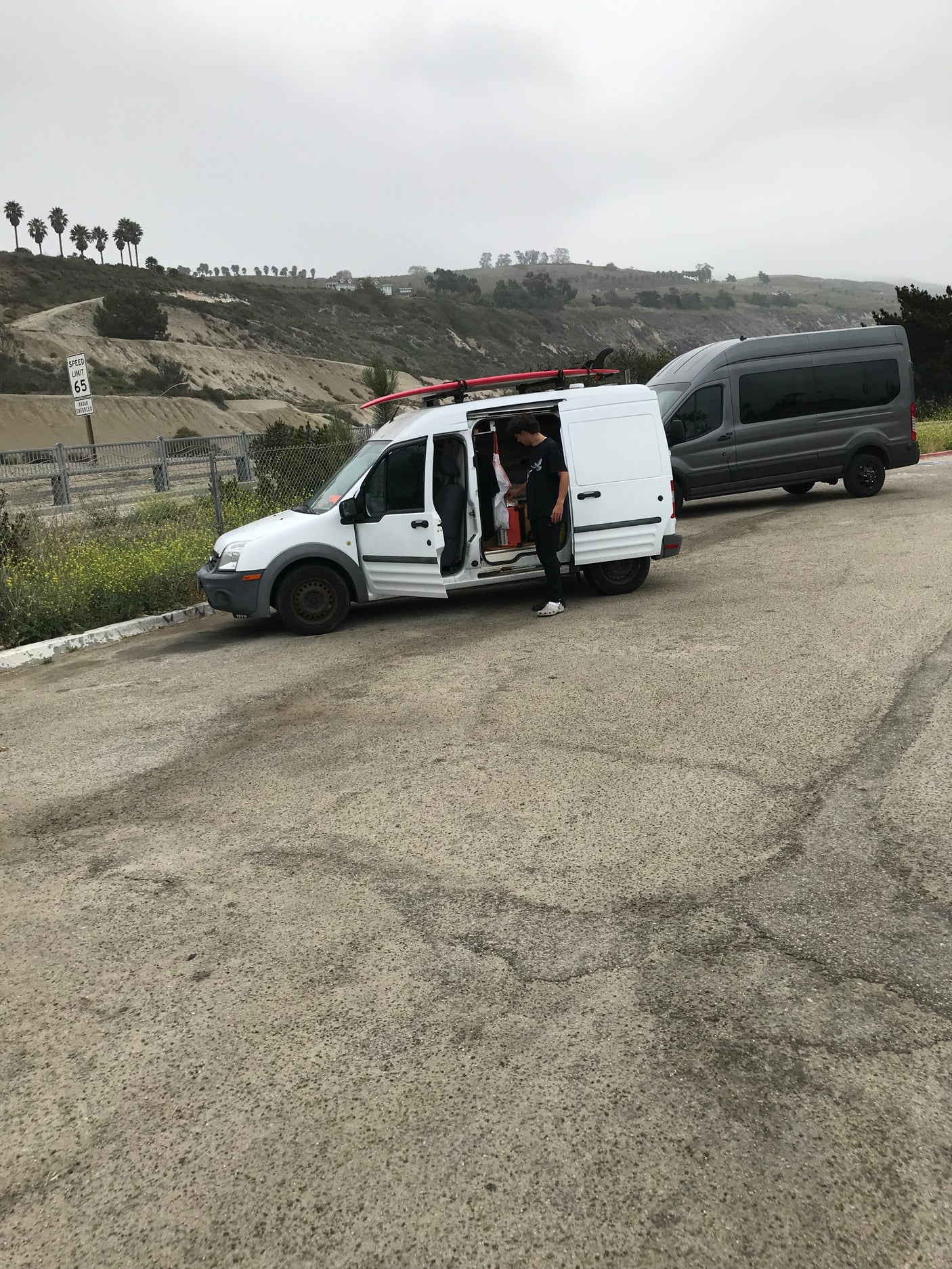 Person organizing inside a white van with a surfboard, alongside a gray van on a scenic hillside. Ideal for an **RV rental** trip.