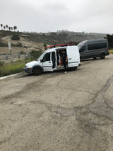 Person organizing inside a white van with a surfboard, alongside a gray van on a scenic hillside. Ideal for an **RV rental** trip.