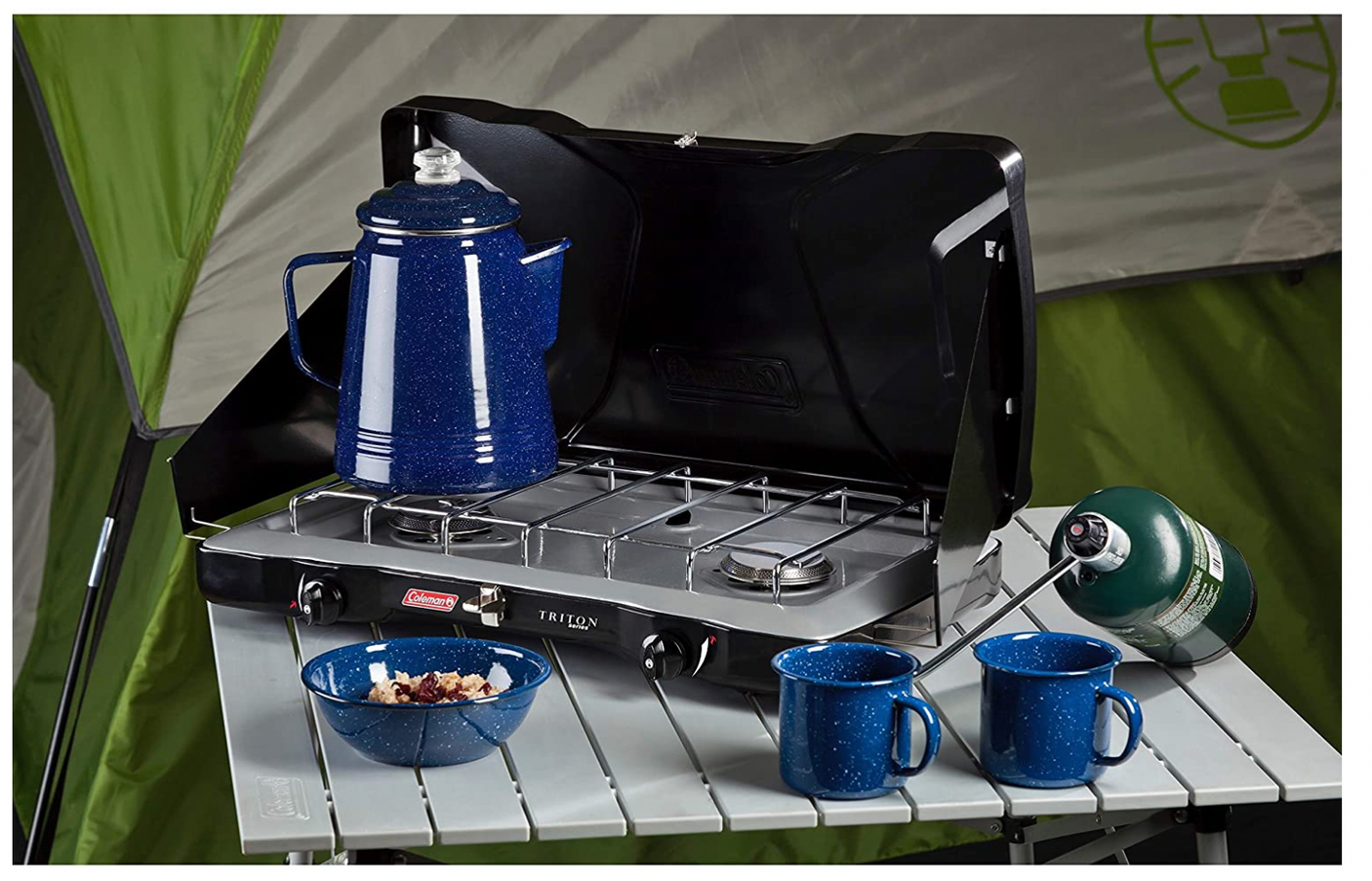 Perfect for RV rental trips: a Coleman camping stove with coffee percolator, mugs, and bowl on a table by a tent.