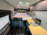 Interior of a modern RV rental camper van, featuring a bed at the rear, a kitchen counter with sink, and passenger seating.