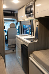 RV rental's galley features a sink, stove, microwave, and fridge, looking towards the driver's cab.