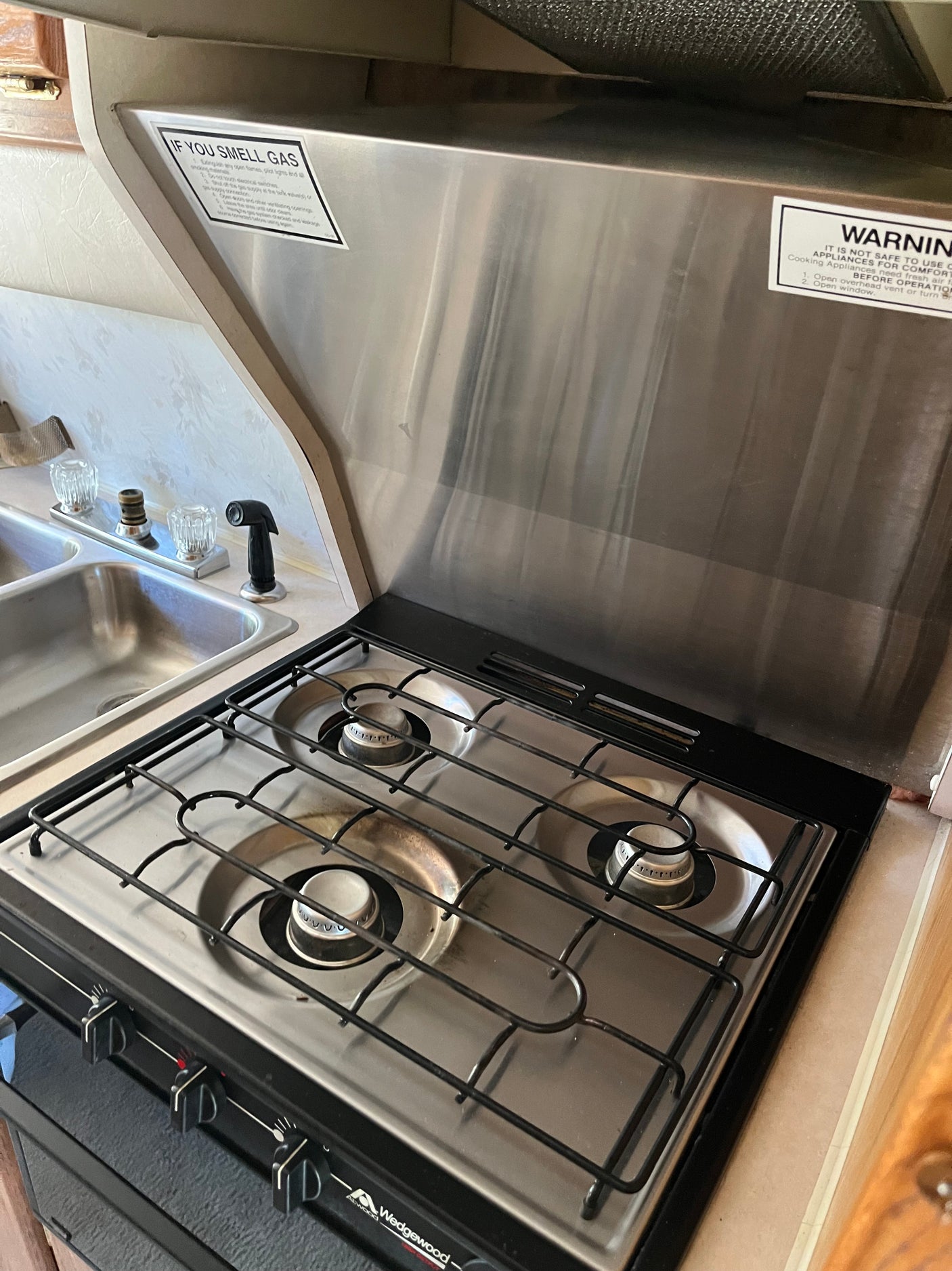 RV rental kitchen features a gas stove with three burners, a metal backsplash, and a double sink. Safety warnings are visible.