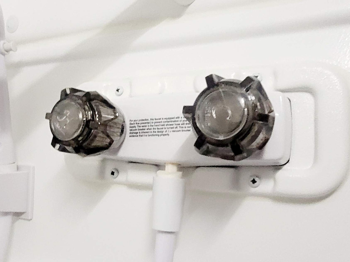 This RV rental's white shower faucet features dual clear handles and a hose connection. Note visible text on backflow prevention.