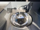 Shiny stainless steel sink & faucet in an rv rental's grey kitchenette. Sunlight highlights the metal & wall textures, casting shadows.