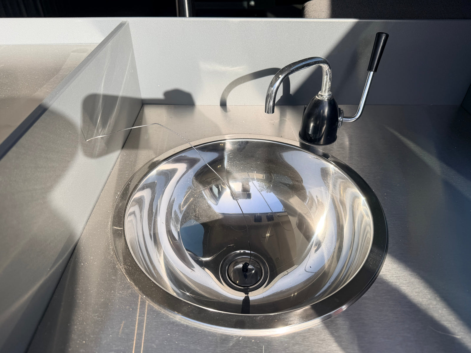 Shiny stainless steel sink & faucet in an rv rental's grey kitchenette. Sunlight highlights the metal & wall textures, casting shadows.