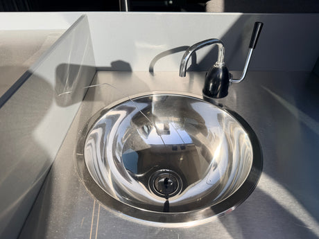 Shiny stainless steel sink & faucet in an rv rental's grey kitchenette. Sunlight highlights the metal & wall textures, casting shadows.