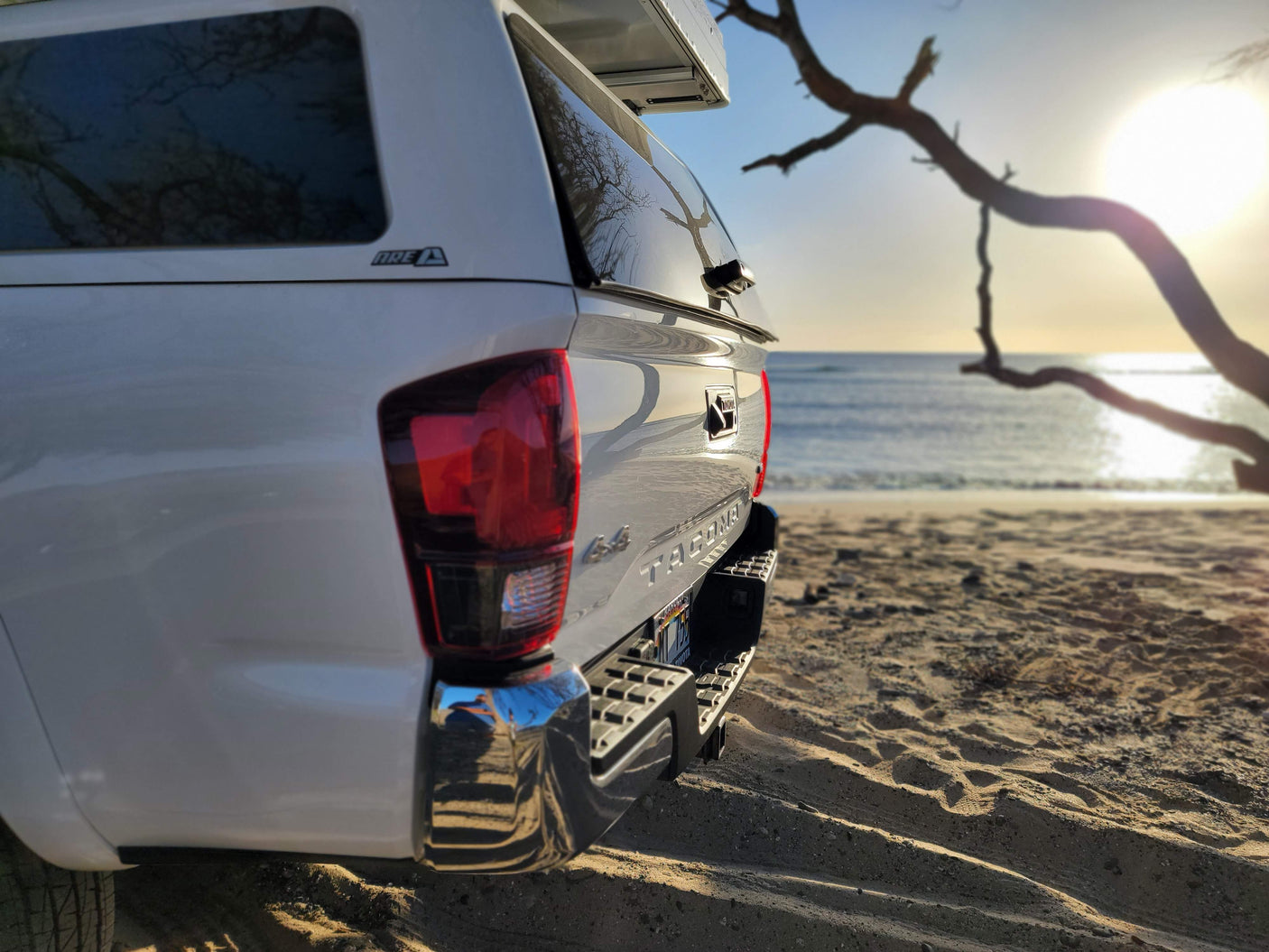 White Toyota Tacoma with camper shell, parked on a sandy beach. An RV rental for rugged outdoor adventures awaits!