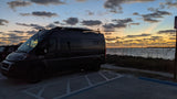 A dark RV rental campervan is parked seaside at sunset, showcasing a beautiful sky. Perfect for travel exploration.
