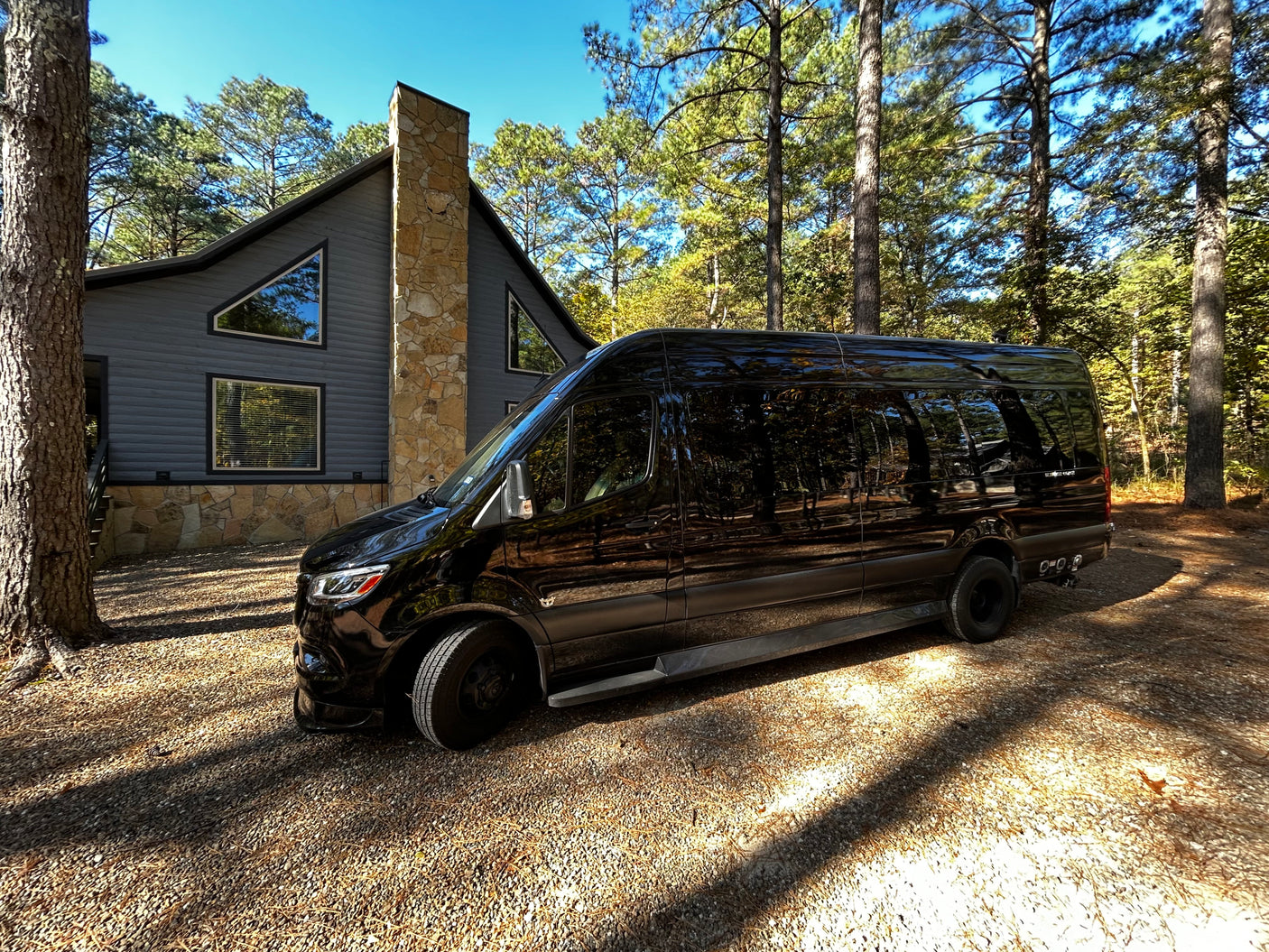 An RV rental, a black Mercedes Sprinter van, parked on a sun-dappled gravel driveway next to a modern cabin in the woods.