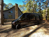 An RV rental, a black Mercedes Sprinter van, parked on a sun-dappled gravel driveway next to a modern cabin in the woods.