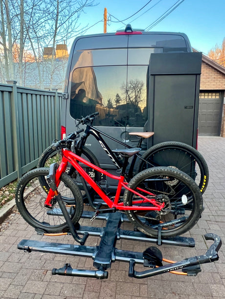 Dark gray RV rental with two mountain bikes (red & black) on a rear hitch rack, plus a storage box, parked on pavers.