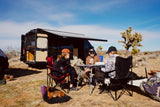 Friends play cards by their **RV rental** in the sunny desert with a dog napping, enjoying van life and a relaxed **camping** trip.