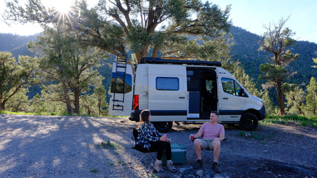 Enjoying nature in an RV rental! A couple relaxes in camping chairs next to their camper van amidst trees and mountains.