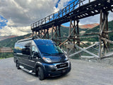Dark RV rental parked at a historic wooden mining structure, with red-streaked mountains under cloudy skies.