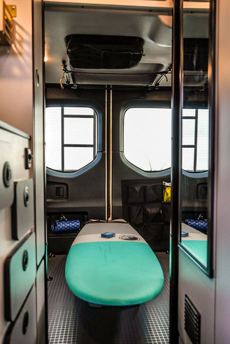 This RV rental's adventure-ready interior showcases a vibrant surfboard centrally, flanked by windows and storage.