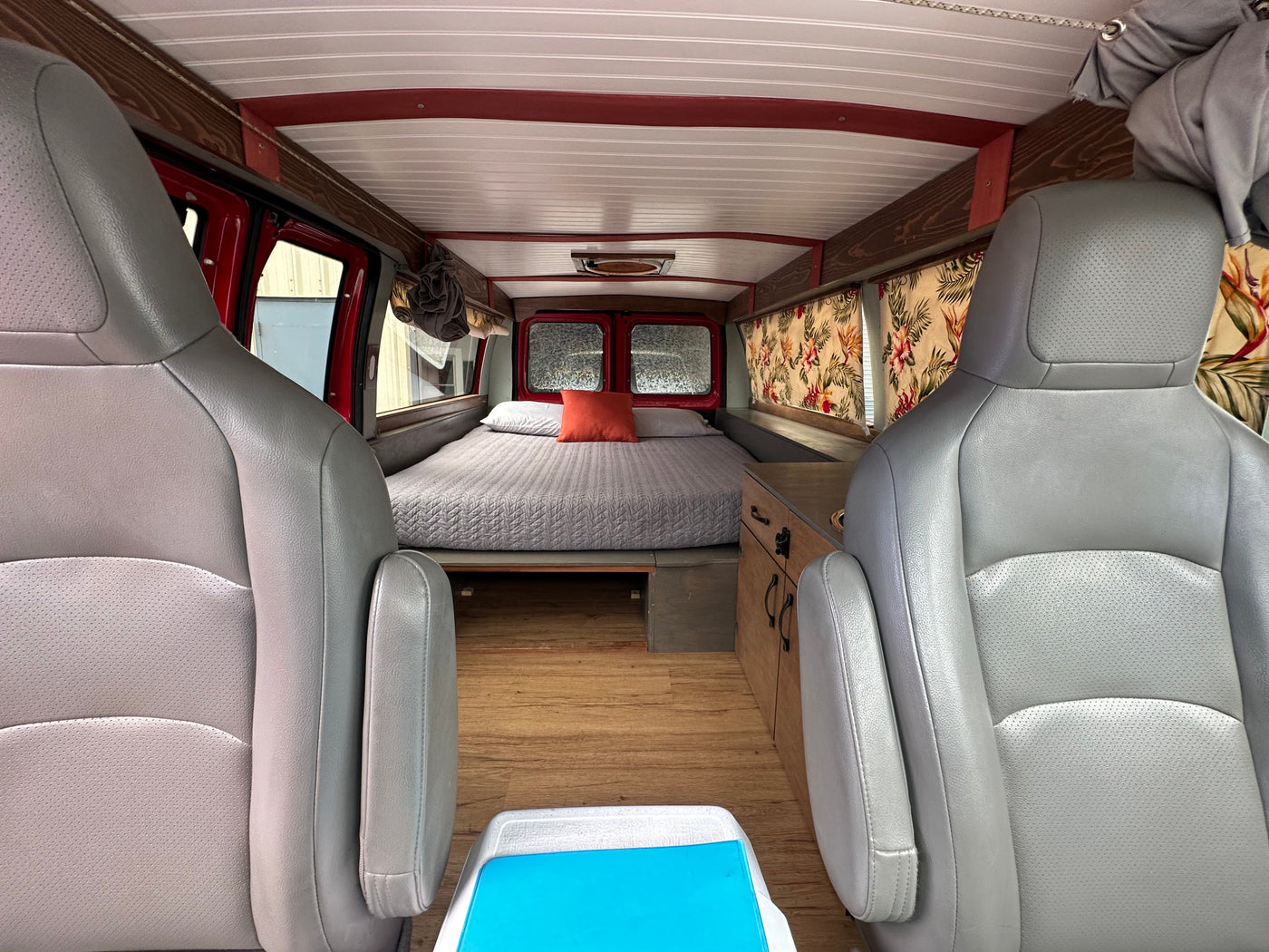 Cozy RV rental interior: grey seats face a comfy bed, wood floors, and a small kitchenette with floral curtains.