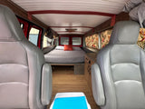Cozy RV rental interior: grey seats face a comfy bed, wood floors, and a small kitchenette with floral curtains.