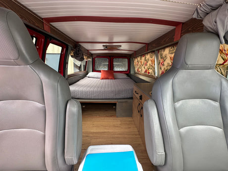 Cozy RV rental interior: grey seats face a comfy bed, wood floors, and a small kitchenette with floral curtains.