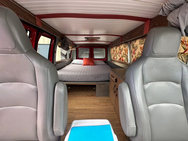 Cozy RV rental interior: grey seats face a comfy bed, wood floors, and a small kitchenette with floral curtains.