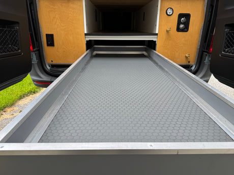 RV rental: Camper van's extended pull-out cargo tray with a textured mat, flanked by wood panels containing a gauge and water connections.