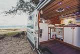 Beachside living: Explore this custom RV rental, a camper van with open door revealing fridge, kitchen, and bed, parked by the ocean.