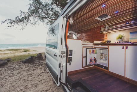 Beachside living: Explore this custom RV rental, a camper van with open door revealing fridge, kitchen, and bed, parked by the ocean.
