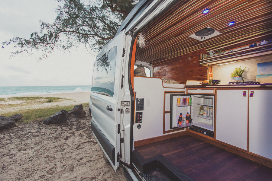 Beachside living: Explore this custom RV rental, a camper van with open door revealing fridge, kitchen, and bed, parked by the ocean.