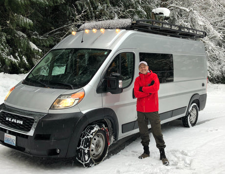 Man next to an RV rental van with tire chains, parked in a snowy forest. Ready for winter adventures!