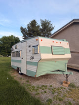 A classic two-tone Shasta camper, ideal for an RV rental, sits on gravel beside a grassy lawn, under a blue sky.
