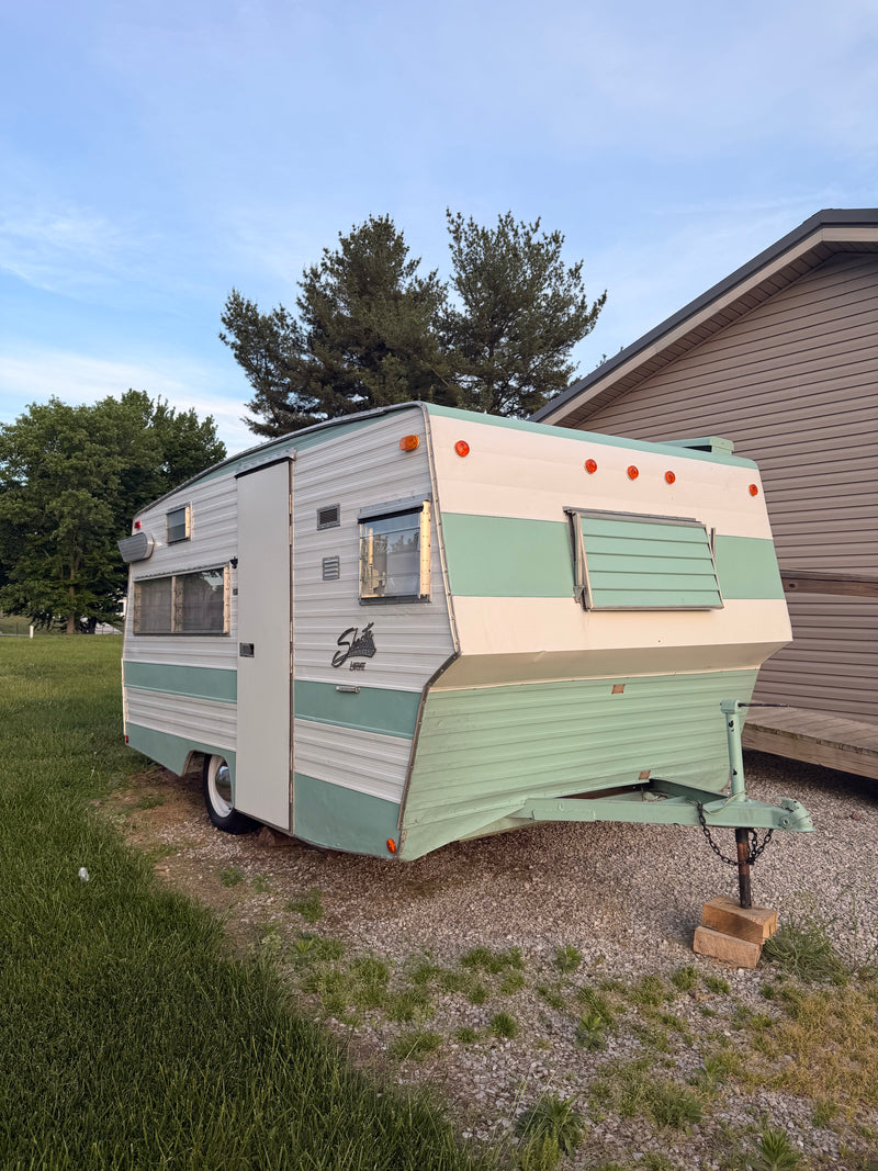 A classic two-tone Shasta camper, ideal for an RV rental, sits on gravel beside a grassy lawn, under a blue sky. — photo 7