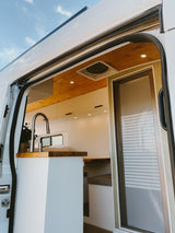 View inside a modern RV rental's bright interior: kitchen, wood ceilings, bed, and frosted sliding door.